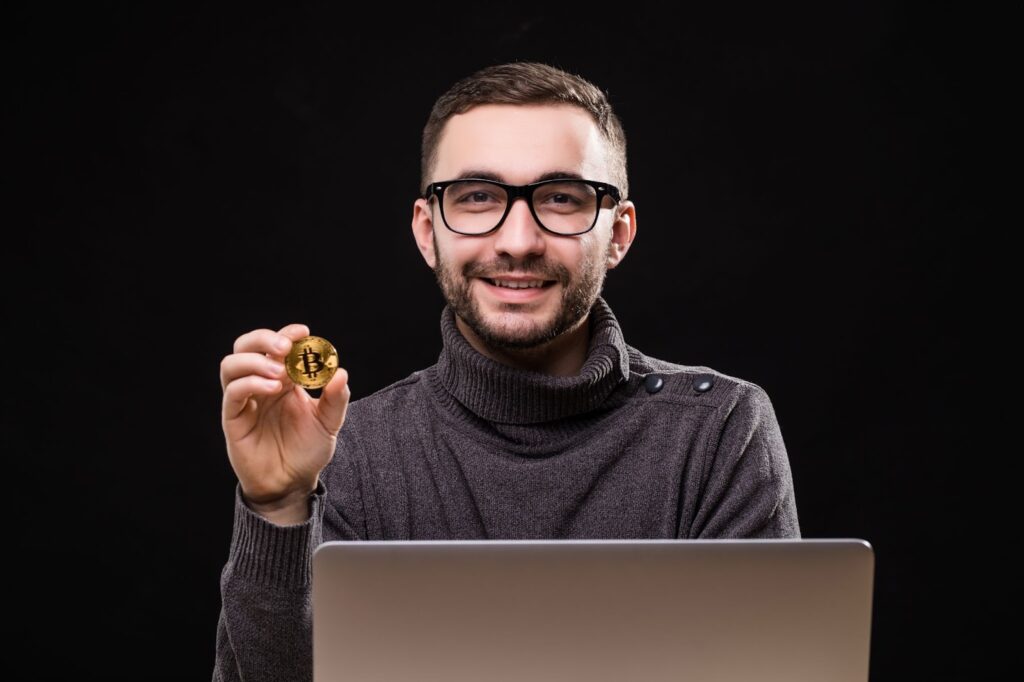 Happy man showing bitcoin while sitting at desk with laptop